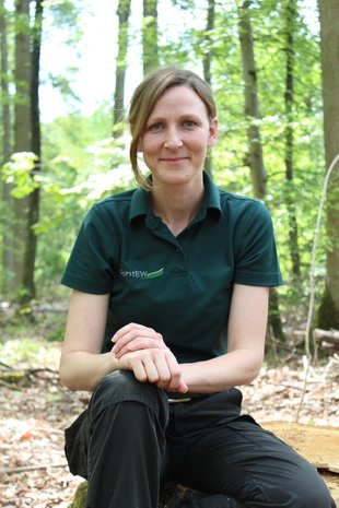 Eine Frau in grünem ForstBW-T-Shirt sitzt im Wald und lächelt in die Kamera.