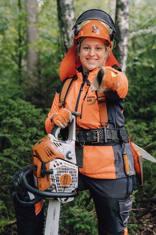 Eine Forstwirt-Auszubildende in Schutzausrüstung und mit einer Kettensäge in der Hand zeigt lächelnd mit ihrem Finger in die Kamera.
