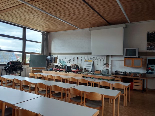 Lehrsaal mit Tischreihen. Vorne sind verschieden Werkzeuge und eine Leinwand an der Wand aufgehängt.
