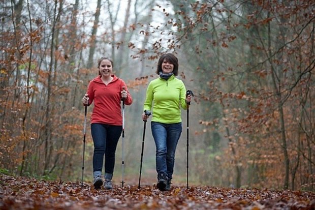 Zwei Frauen, die mit Nordic-Walking-Stöcken in einer herbstlichen Umgebung auf die Kamera zulaufen.